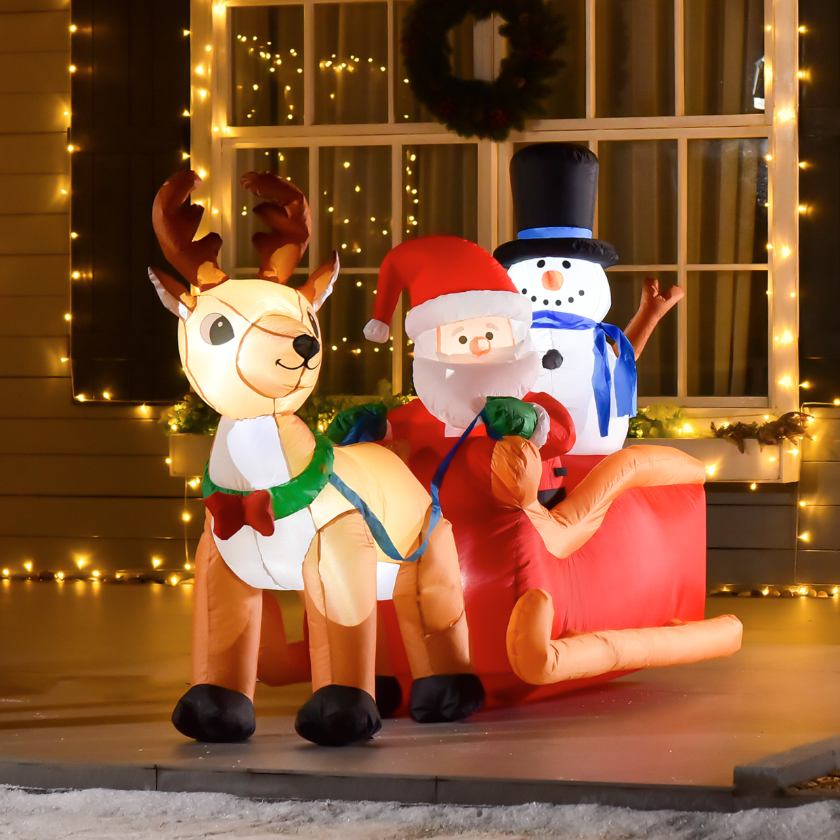 Inflatable reindeer and Santa Claus with a snowman on a sled, decorated for Christmas, in front of a window with lights.